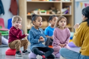 A small group of preschool children sit on the floor in front f their teacher as she leads them in a song.  The children are singing along and doing the actions as they follow the teachers lead.