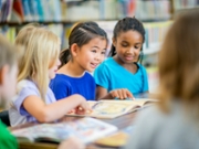 A group of elementary school girls of different ethnicity are indoors in their school library. They are sitting together at a table and reading together.