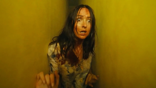A disheveled and frightened looking young woman with long dark hair in the corner of a yellow-walled room