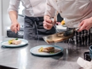 Cook in uniform putting garnish on plate from pan with chef standing behind him and controlling the process