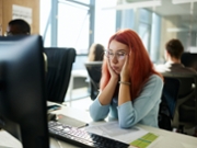 Young redhead female designer feeling exhausted while working on desktop computer in the office.