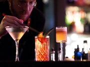 A still photograph of a bartender as he puts an orange slice into a cocktail. The drinks are vibrant and brightly lit, standing out against the low-key lighting of the background. The drinks and bartender's hand are in focus, while the rest remains blurry.