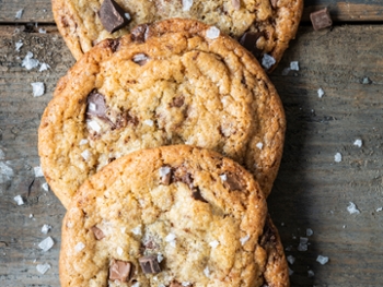 Three freshly baked chocolate chip cookies are lying on a textured wooden background sprinkled with salt. There are loose chocolate chips and salt flakes scattered around the cookies. The chocolate in the cookies has melted leaving the biscuits gooey. Taken from overhead.
