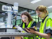 Two women wearing safety vests and goggles are looking at a robot. One of them is holding a tablet