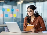 Woman watching online video course, training conference inside office at workplace. Office worker in headphones using laptop for remote meeting, writing data in notebook.