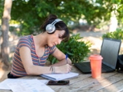 University student is taking notes at a picnic table on campus while listening to music on her headphones