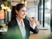 A confident young businesswoman, dressed professionally, smiles as she efficiently uses voice commands on her smartphone in a modern office or cafe setting. This image perfectly captures the essence of voice technology in contemporary business, highlighting its crucial role in enhancing mobile productivity, streamlining communication, and driving efficiency for busy professionals in a digitally transformed world