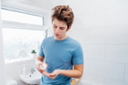 Concentrated caucasian teenage boy with acne problem takes care his face skin at home. He squeezing washing lotion for cleaning face in bathroom. Teenager skin care cosmetics. Selective focus