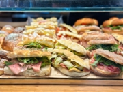 Close-up of multiple rustic sandwiches displayed on a bakery counter, featuring crusty bread filled with a mix of cured meats, arugula, and grilled vegetables. The arrangement emphasizes the textures of the bread, the vivid green of the leafy vegetables, and the marbled tones of the sliced meats. Natural lighting enhances the freshness and appetizing appearance of the sandwiches.