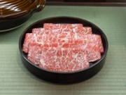 霜降り黒毛和牛 A top-down, close-up shot of a dark brown, shallow platter containing thinly sliced, raw Wagyu beef. The beef is a vibrant red with extensive white marbling, characteristic of high-quality Wagyu. The slices are neatly folded and arranged in a single layer to show off the marbling. The platter rests on a light green tatami mat, suggesting the beef is prepared for a traditional Japanese hot pot meal (nabe).