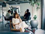 Non-binary person takes a mental health break in a modern office with incidental people in the background.
