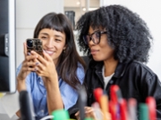 Women coworkers taking a break from work, sharing a fun moment using a phone together in a modern office space.