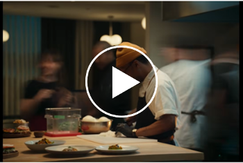 Male standing at an island counter slicing onions in a restaurant's kitchen with blurry views of other employees around him.