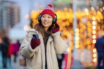 A cheerful woman wearing headphones standing outside near festive lights, holding her phone.