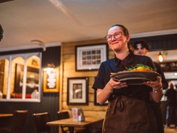 Young waitress carrying and serving a gourmet restaurant meal in a cosy gastropub dining room. Authentic hospitality and customer service scene capturing food delivery, casual dining experience and warm restaurant atmosphere in a modern British pub restaurant.