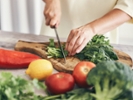 A close-up shot of hands chopping leafy greens on a wooden cutting board in a kitchen setting. Fresh vegetables such as a red bell pepper, lemon, and tomatoes are scattered around the cutting board. The scene depicts a vibrant and healthy food preparation process with a focus on fresh ingredients.