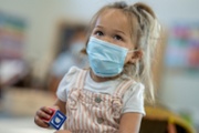 Cute little girl wearing a medical mask while sitting on the carpet in a daycare facility.