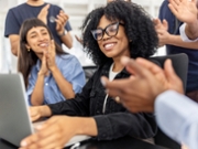 African woman sitting at desk shares good news with her team clapping and celebrating a business achievement in a lively startup office. Excited team celebrates good news with colleague at desk in startup office.