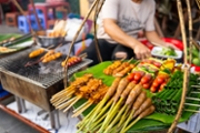 A vibrant street food stall showcasing a variety of grilled food skewers and fresh ingredients, artistically presented on banana leaves, representing local culinary traditions and culture in an outside urban setting.