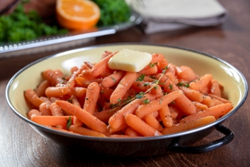 Serving dish of honey glazed baby carrots seasoned with salt, pepper, thyme and butter. It sits on a table with dinner in the background out of focus.