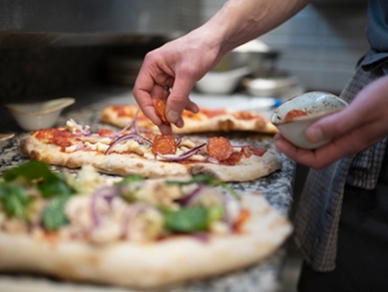 Chef placing sausage onto Pinsa Romana base, a Roman style pizza blend reducing sugar and saturated fat, containing rice and soy with less gluten, close up of hands