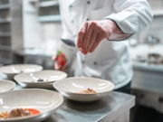 A professional chef in a modern kitchen carefully garnishes plates with precision and creativity. The setting features polished countertops, white chef attire, and a focus on culinary artistry and preparation.