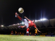 Wide shot low angle still of a young adult professional football player skillfully jumping up and kicking a soccer ball bicycle kick style in mid-air while illuminated by stadium floodlights during an international football match