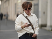 MILAN, ITALY - JANUARY 18: A guest wears short tousled light brown hair with a middle part, small oval dark-lens sunglasses with thin gold metal frames, a gold metal watch and a gold ring, a brown canvas crossbody bag with leather trim and a brass buckle, a cream knit sweater with a folded collar and ribbed cuffs tucked into dark indigo blue denim jeans pants with a dark brown leather belt and silver buckle, shoes, outside Prada, during Milan Fashion Week - Menswear Fall/Winter 2026-2027, on January 18, 2026 in Milan, Italy (Photo by Edward Berthelot/Getty Images)