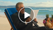 Older man sitting on a lounge couch, facing left to right, on a beach reading a piece of paper.