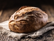 An artisan loaf of bread with a golden brown, crispy crust. The loaf is placed on a crumpled, light-colored linen towel that rests on a rustic wooden table with a dark background.