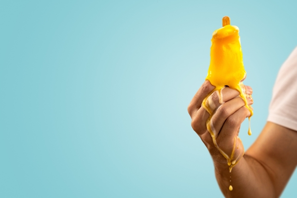 A hand holding a yellow ice pop that's rapidly melting. Isolated on a light blue background with copy space on the left.