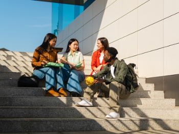 Diverse young adult friends sitting on concrete stairs, talking and smiling while holding books on a college campus
