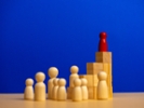 Red wooden figure standing on top of a structure made of wooden blocks, leading a group of uncolored wooden figures, symbolizing leadership, success, and achievement