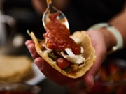 A vibrant scene of a hand adding salsa onto a taco, filled with colorful vegetables and toppings. Perfect for food enthusiasts, cooking events, and recipe presentations.